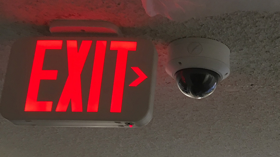 CCTV dome camera mounted on a facility ceiling near an exit sign